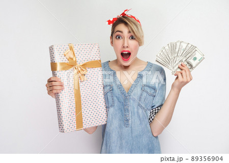Portrait of amazed young girl in casual denim shirt with makeup and red headband standing, showing gift box and fan dollars with opened mouth. indoor studio shot, isolated on white background 89356904