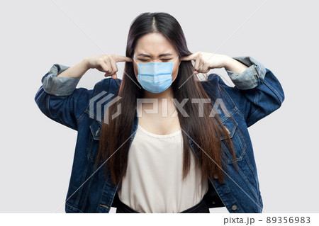 Portrait of nervous brunette asian young woman with surgical medical mask in casual blue jacket standing and putting finger on ears and don't want to hear. studio shot, isolated on grey background. Portrait of nervous brunette asian young woman with surgical medical mask in casual blue jacket standing and putting finger on ears and don't want to hear. studio shot, isolated on grey background. 89356983