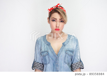 Portrait of sad beautiful young woman in casual blue denim shirt with makeup and red headband standing and looking at camera with unhappy face. indoor studio shot, isolated on white background. Portrait of sad beautiful young woman in casual blue denim shirt with makeup and red headband standing and looking at camera with unhappy face. indoor studio shot, isolated on white background. 89356984