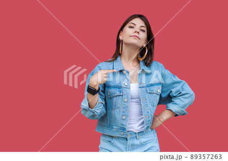 Portrait of proud beautiful brunette young woman with makeup in denim casual style standing, pointing herself and looking at camera with haughty face. indoor studio shot, isolated on red background. 89357263