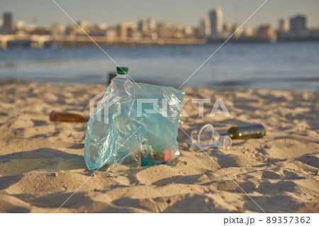 Blue bag with garbage lies on beach of picturesque river, crumpled plastic and glass bottles are scattered nearby. Cleaning up trash. Close-up. Blue bag with garbage lies on beach of picturesque river, crumpled plastic and glass bottles are scattered nearby. Cleaning up trash. Close-up. 89357362