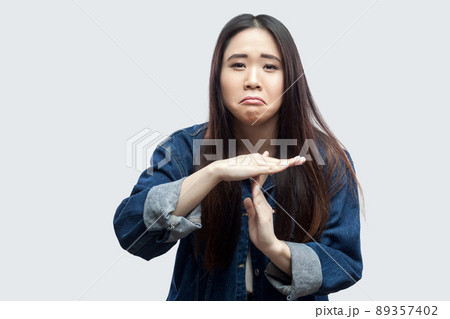 Portrait sad of beautiful brunette asian young woman in casual blue denim jacket with makeup standing and looking at camera with timeout sign. indoor studio shot, isolated on light grey background. Portrait sad of beautiful brunette asian young woman in casual blue denim jacket with makeup standing and looking at camera with timeout sign. indoor studio shot, isolated on light grey background. 89357402