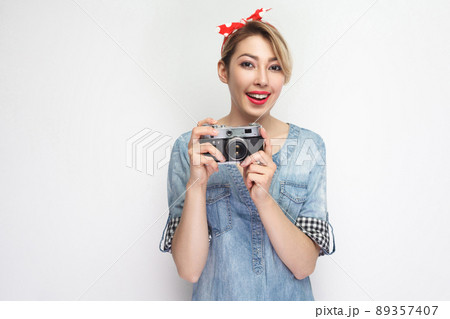 Portrait of beautiful young blogger woman in casual blue denim shirt with makeup and red headband standing, holding retro camera with toothy smile. indoor studio shot, isolated on white background. 89357407