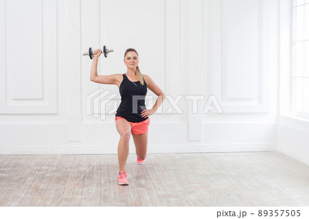 Portrait of young athletic beautiful bodybuilder woman in pink shorts and black top doing squats and exercising at the gym with the dumbells on white wall. indoor, studio shot, looking at camera 89357505