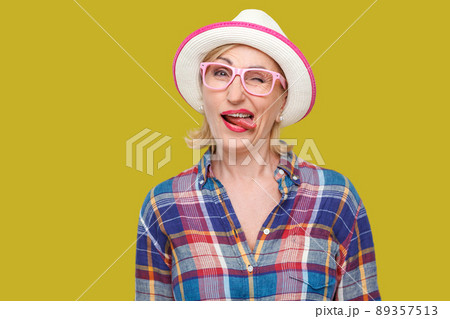 Portrait of funny crazy modern stylish mature woman in casual style with hat and eyeglasses standing, winking, tongue out and looking at camera. indoor studio shot isolated on yellow background. Portrait of funny crazy modern stylish mature woman in casual style with hat and eyeglasses standing, winking, tongue out and looking at camera. indoor studio shot isolated on yellow background. 89357513