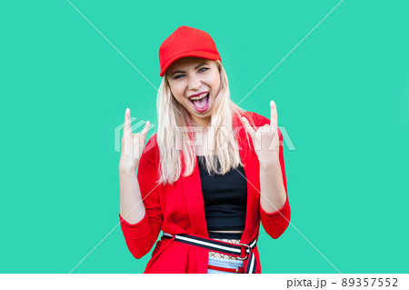 Portrait of happy funny amazed beautiful blond young hipster style woman in red blouse and cap, standing with rock gesture sign and looking at camera. indoor studio shot, isolated on green background. 89357552
