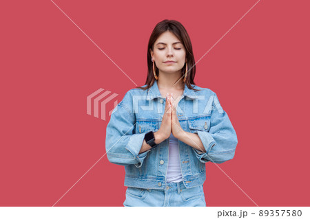 Portrait of calm beautiful brunette young woman with makeup in denim casual style standing with palm hands and doing meditating in yoga pose. indoor studio shot, isolated on red background. 89357580