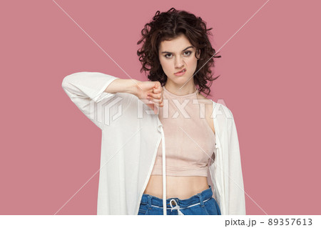 Dislike. Portrait of dissatisfied beautiful brunette young woman with curly hairstyle in casual style standing, looking at camera, holding thumbs down. indoor studio shot isolated on pink background. Dislike. Portrait of dissatisfied beautiful brunette young woman with curly hairstyle in casual style standing, looking at camera, holding thumbs down. indoor studio shot isolated on pink background. 89357613