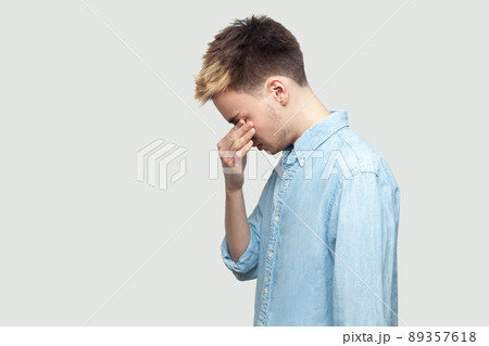Profile side view portrait of depressed sad handsome young man in light blue shirt standing, covering his eyes and crying. indoor studio shot on grey background copy space. Profile side view portrait of depressed sad handsome young man in light blue shirt standing, covering his eyes and crying. indoor studio shot on grey background copy space. 89357618