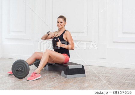 Happy beautiful young athletic causcasian woman pink shorts and black top siting, drink water and have a rest after hard working on the gym. Indoor, studio shot, sport concept, looking at camera 89357619