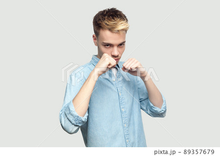 Portrait of serious handsome young man in light blue shirt standing in boxing fists, looking at camera and ready to attack or defence. indoor studio shot on grey background copy space. Portrait of serious handsome young man in light blue shirt standing in boxing fists, looking at camera and ready to attack or defence. indoor studio shot on grey background copy space. 89357679