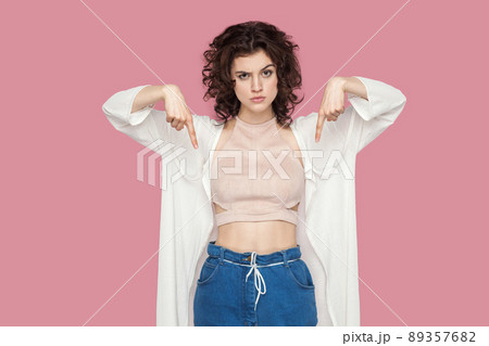 Here and right now. Portrait of serious cute brunette young woman with curly hairstyle in casual style standing and pointing down and looking at camera. indoor studio shot isolated on pink background. 89357682