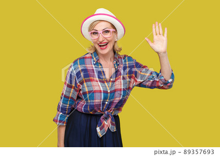 Portrait of surprised modern stylish mature woman in casual style with hat and eyeglasses standing waving her hand and looking at camera with toothy smile. studio shot isolated on yellow background. Portrait of surprised modern stylish mature woman in casual style with hat and eyeglasses standing waving her hand and looking at camera with toothy smile. studio shot isolated on yellow background. 89357693