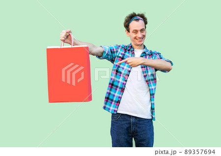 Cheerful modern young man in white t-shirt and checkered shirt standing and pointing finger to shopping bags and toothy smile, looking at camera. Studio shot, green background, isolated, indoor 89357694