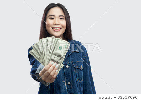 Portrait of successful attractive young asian girl in blue denim jacket standing, holding many dollars, demonstration and looking at camera with smile. Indoor, studio shot, isolated, gray background 89357696