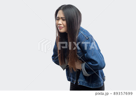 Stomach pain. Side view portrait of beautiful brunette asian young woman in casual blue denim jacket standing and feeling pain on her stomach. indoor studio shot, isolated on light grey background. 89357699