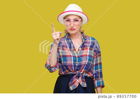 Portrait of serious bossy modern stylish mature woman in casual style with hat and eyeglasses standing with warn sign and looking at camera. indoor studio shot isolated on yellow background. 89357700