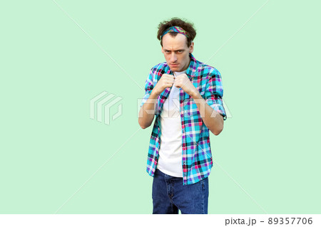 Portrait of angry young man in casual blue checkered shirt and headband standing and looking at camera with boxing fists and ready to attack. indoor studio shot, isolated on light green background. 89357706