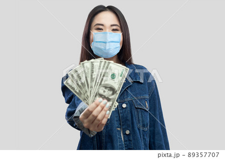 Portrait of successful attractive young asian woman with medical mask standing, holding dollars, demonstration and looking at camera with smile. Indoor, studio shot, isolated, gray background 89357707