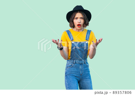 Portrait of shocked pretty young hipster girl in blue denim overalls, yellow shirt and black hat standing with raised arms and looking at camera. indoor studio shot isolated on light green background. Portrait of shocked pretty young hipster girl in blue denim overalls, yellow shirt and black hat standing with raised arms and looking at camera. indoor studio shot isolated on light green background. 89357806