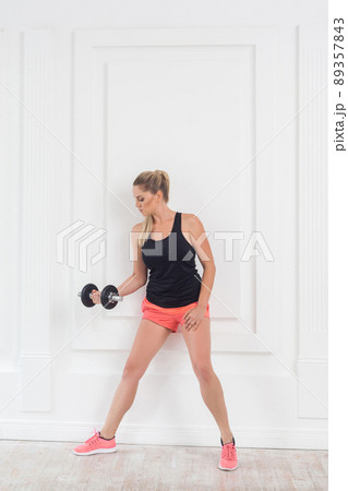 Young adult motivation athletic beautiful bodybuilder woman in pink shorts and black top standing, holding dumbells doing exercise for arms at the gym, lean on white wall. indoor, studio shot Young adult motivation athletic beautiful bodybuilder woman in pink shorts and black top standing, holding dumbells doing exercise for arms at the gym, lean on white wall. indoor, studio shot 89357843