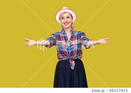 Portrait of happy peaceful modern stylish mature woman in casual style with white hat standing with raised arms, looking at camera with toothy smile. indoor studio shot isolated on yellow background. 89357923