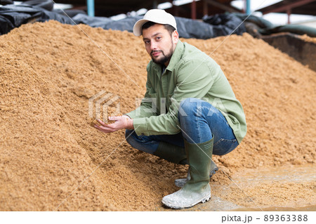 Farmer squatting at large heap of brewer's spent grain Farmer squatting at large heap of brewer's spent grain 89363388