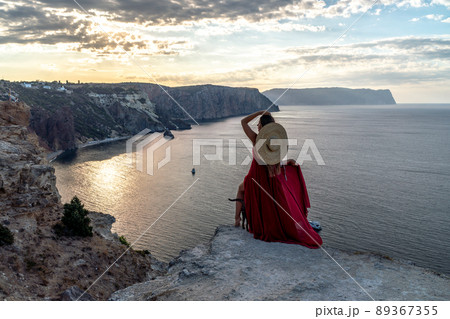 A girl with loose hair in a red dress and hat stands on the rocks above the sea. In the background, the sea and the rocks. The concept of travel. 89367355