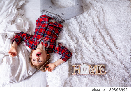 Funny little curly kid boy with smiley face and open mouth lying on the white bed at home. Stay at home quarantine coronavirus COVID-19 pandemic prevention. With space for text and wooden house Funny little curly kid boy with smiley face and open mouth lying on the white bed at home. Stay at home quarantine coronavirus COVID-19 pandemic prevention. With space for text and wooden house 89368995