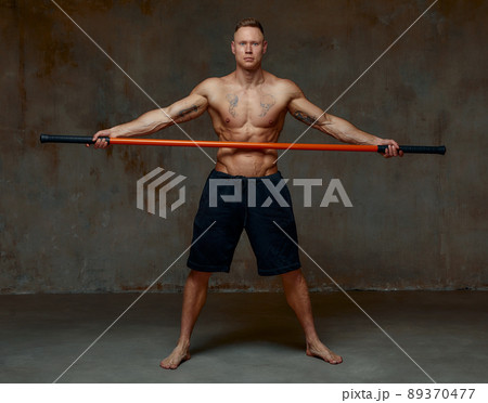 A fighter shirtless in black shorts, stands in a fighting artificially pose and holds a fight stick on studio background. A fighter shirtless in black shorts, stands in a fighting artificially pose and holds a fight stick on studio background. 89370477