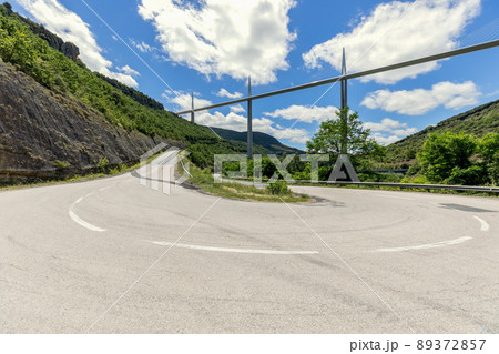 Flat straight Millau Viaduct above and old curly country road D41 on ground beneath 89372857