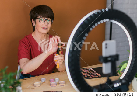 Middle-aged female beauty blogger or influencer with makeup product recording video in room at home. Using ring lamp and smartphone Middle-aged female beauty blogger or influencer with makeup product recording video in room at home. Using ring lamp and smartphone 89379711
