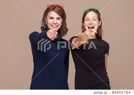 Women standing near each other and pointing fingers at camera. Studio shot, light brown background Women standing near each other and pointing fingers at camera. Studio shot, light brown background 89405764