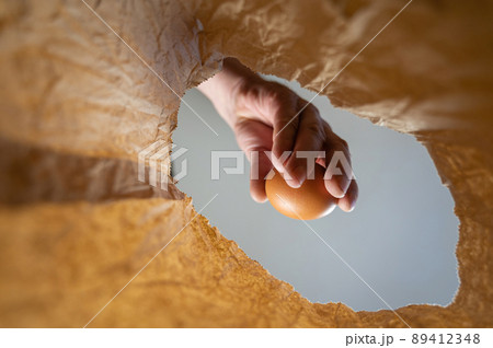 A hand takes a chicken egg out of a paper bag. A mature man's hand holds a brown egg over an open brown bag. Shot from the bottom up. Close up. Selective focus A hand takes a chicken egg out of a paper bag. A mature man's hand holds a brown egg over an open brown bag. Shot from the bottom up. Close up. Selective focus 89412348