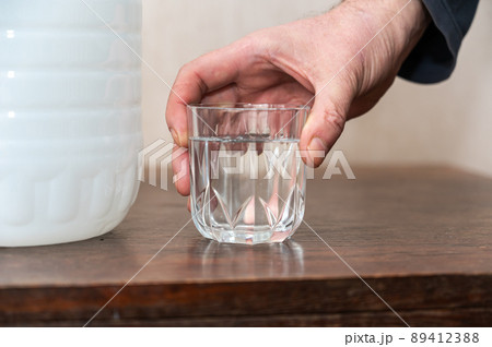 A man holds a glass of drinking water in his hand. A large white plastic bottle of fresh water is next to him on the table. Inside the room. A man holds a glass of drinking water in his hand. A large white plastic bottle of fresh water is next to him on the table. Inside the room. 89412388