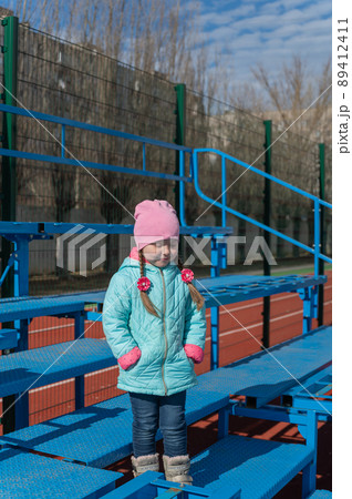 Full-length portrait of a five-year-old girl in an empty sports stand. The child stands on the blue tribune of the school stadium holding her hands in her jacket pockets. Full-length portrait of a five-year-old girl in an empty sports stand. The child stands on the blue tribune of the school stadium holding her hands in her jacket pockets. 89412411
