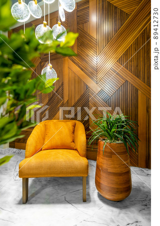 Modern orange armchair, tall rounded wooden planter with green bushes, tall glass chandelier, and wood cladding wall Modern orange armchair, tall rounded wooden planter with green bushes, tall glass chandelier, and wood cladding wall 89412730