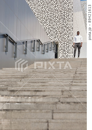 Athletic Black man in stylish sportswear standing on concrete stairs against contemporary office building with perforated facade, low angle view Athletic Black man in stylish sportswear standing on concrete stairs against contemporary office building with perforated facade, low angle view 89421613