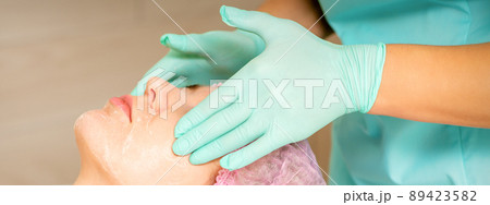 Cosmetologist with gloved hands applies a moisturizing mask with peeling cream on the female face. Facial cosmetology treatment. Procedures for facial care Cosmetologist with gloved hands applies a moisturizing mask with peeling cream on the female face. Facial cosmetology treatment. Procedures for facial care 89423582