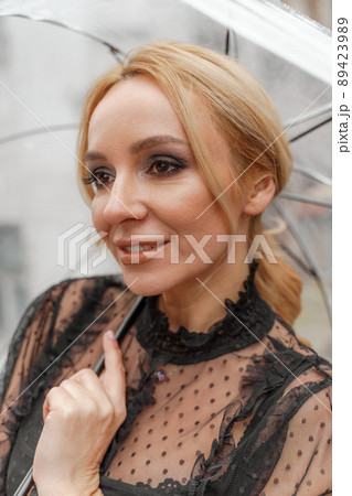 The blonde stands under a transparent umbrella during the rain. The fall season. Rear view. The woman is dressed in a black lace dress, her hair pulled back in a ponytail. 89423989