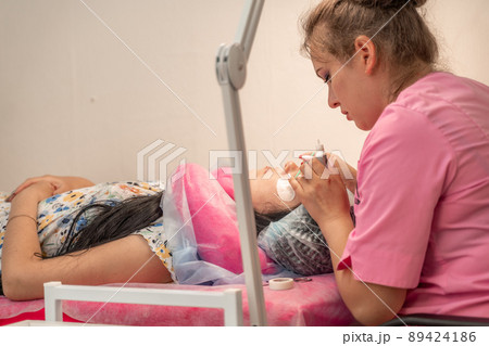 Master lashmaker, diligently extends eyelashes to the client, on the couch, in the beauty salon. A beauty master, a girl of 30 years old, European appearance, at work, in a bright room 89424186