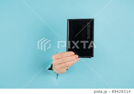 A woman's hand sticks out of a hole in a blue paper background and holds a passport in a black cover. A woman's hand sticks out of a hole in a blue paper background and holds a passport in a black cover. 89426314