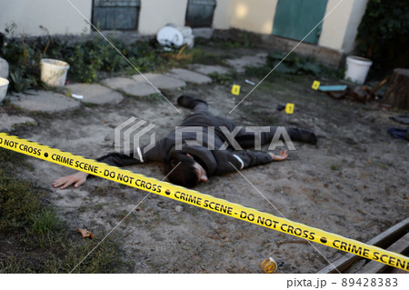 Victim of a violent crime in a backyard of residental house in evening. Dead man body under the yellow police line tape and evidence markers on crime scene 89428383