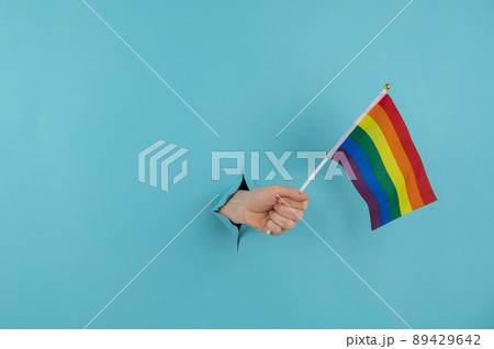A woman's hand sticks out of a hole in a blue paper background and holds an lgbt flag. A woman's hand sticks out of a hole in a blue paper background and holds an lgbt flag. 89429642