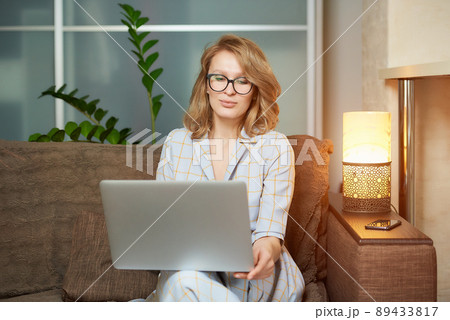 A young woman in glasses works remotely on a laptop in her apartment. 89433817