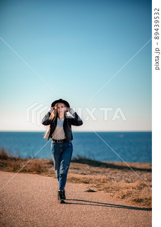 A blonde in a stylish black leather jacket walks along the seashore. 89439532