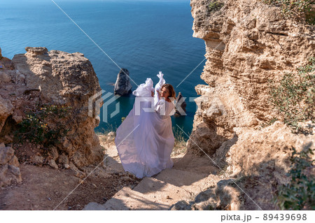 A beautiful young woman in a white light dress with long legs stands on the edge of a cliff above the sea waving a white long dress, against the background of the blue sky and the sea. A beautiful young woman in a white light dress with long legs stands on the edge of a cliff above the sea waving a white long dress, against the background of the blue sky and the sea. 89439698