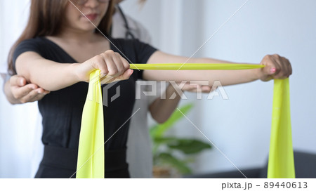 Female patient stretching yellow elastic band expander for rehabilitation exercises under physiotherapist supervision. 89440613