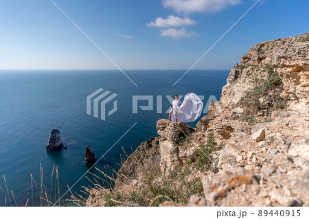 A beautiful young woman in a white light dress with long legs stands on the edge of a cliff above the sea waving a white long dress, against the background of the blue sky and the sea. A beautiful young woman in a white light dress with long legs stands on the edge of a cliff above the sea waving a white long dress, against the background of the blue sky and the sea. 89440915