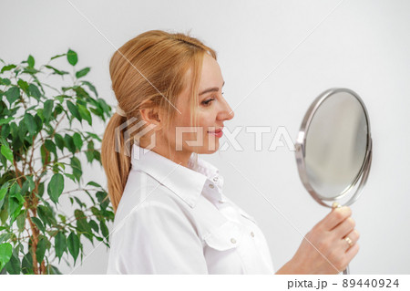 A blonde woman in white formal clothes looks at herself in the mirror. On a white background, green leaves at the back. A blonde woman in white formal clothes looks at herself in the mirror. On a white background, green leaves at the back. 89440924